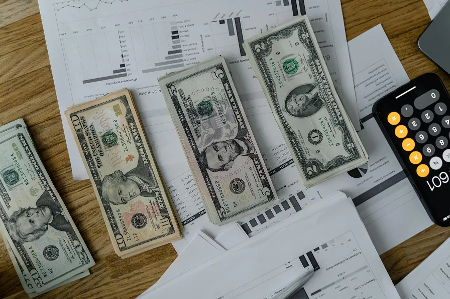 A desk with stacks of U.S. dollar bills, a calculator, and financial charts, representing the financial planning and tracking of FSA or HSA healthcare savings.