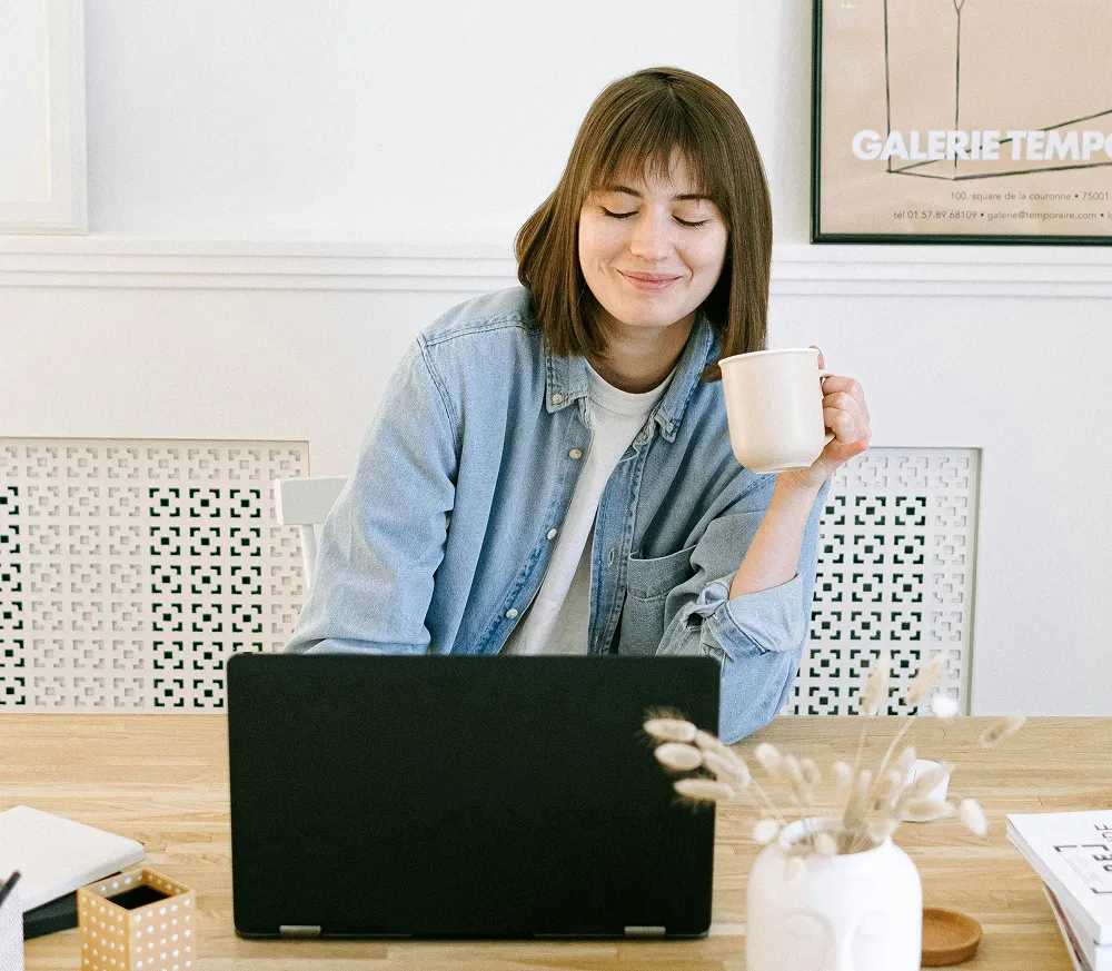A woman in a blue denim shirt sipping coffee while working on a laptop in a cozy office, symbolizing a relaxed approach to managing FSA or HSA healthcare plans.