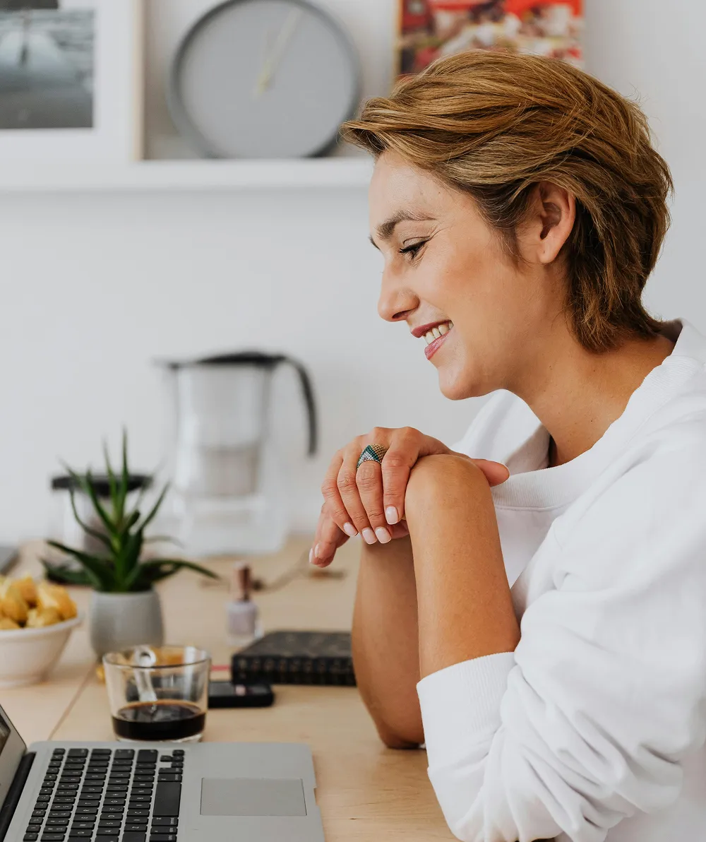 A woman with short hair sitting at a desk with a laptop, smiling thoughtfully while managing her FSA or HSA account details in a cozy home office setting.