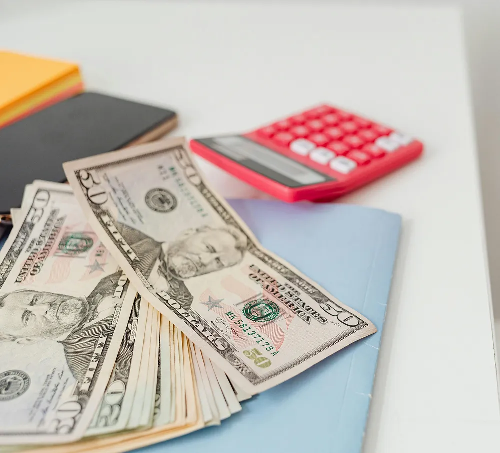 A stack of U.S. dollar bills on a desk next to a red calculator and notebooks, symbolizing the budgeting and calculation process for FSA or HSA healthcare funds.